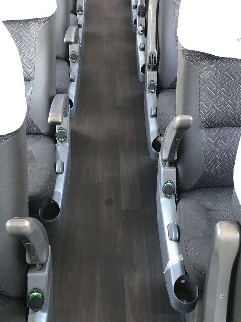 Interior de autobús con filas de asientos de tela gris estampada, suelo de madera oscura y tomas de carga USB visibles con luz indicadora verde en los reposabrazos.